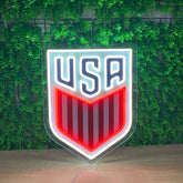 A shield-shaped USA soccer logo sign with a glowing white border, navy blue 'USA' lettering, and red and white vertical stripes, displayed against a green leaf wall.