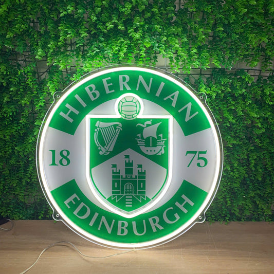 A circular illuminated Hibernian Edinburgh sign with a green and white design, displayed on a wooden surface against a green ivy wall background.
