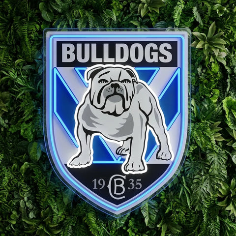 A shield-shaped bulldog emblem with the word BULLDOGS at the top, a gray bulldog illustration in the center, and blue geometric accents, shown against a green leafy background.