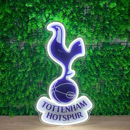 A Tottenham Hotspur illuminated LED sign featuring the club's cocker and football logo in purple and white, set against a green leaf wall background.