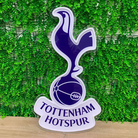 A glossy Tottenham Hotspur club crest sign featuring the team's cocker and football emblem in dark blue with a white border, mounted on a white scroll reading 'TOTTENHAM HOTSPUR', set against a green artificial ivy wall background.
