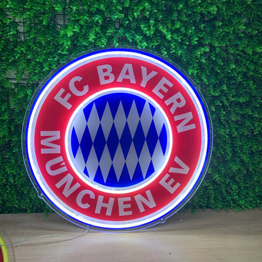 A circular illuminated FC Bayern Munich LED neon sign displayed against a green artificial ivy wall backdrop.