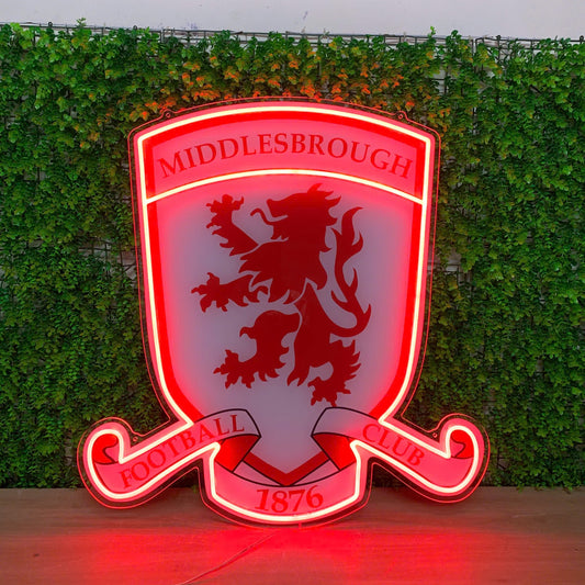 A red and white illuminated LED neon sign of the Middlesbrough Football Club crest, displayed on a wooden surface against a green ivy wall background.