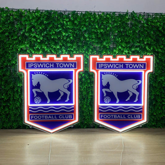 Two illuminated Ipswich Town Football Club neon-style wall signs displayed side by side in front of a green artificial foliage backdrop.