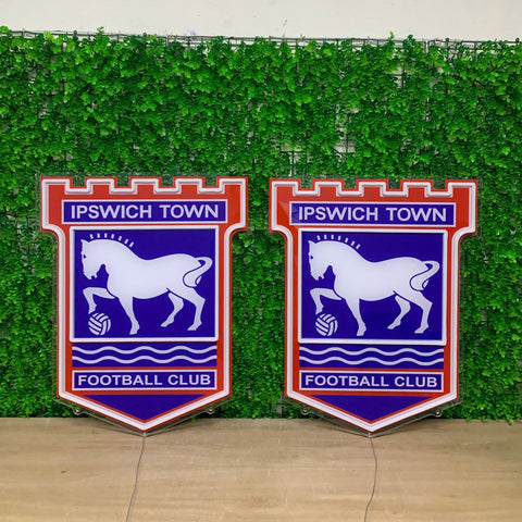 Two matching Ipswich Town Football Club shield-shaped wall signs displayed side by side in front of a green artificial hedge wall.