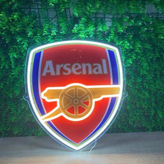 A glowing Arsenal football club shield sign with a red, blue, and gold cannon emblem, set against a green leaf wall background.