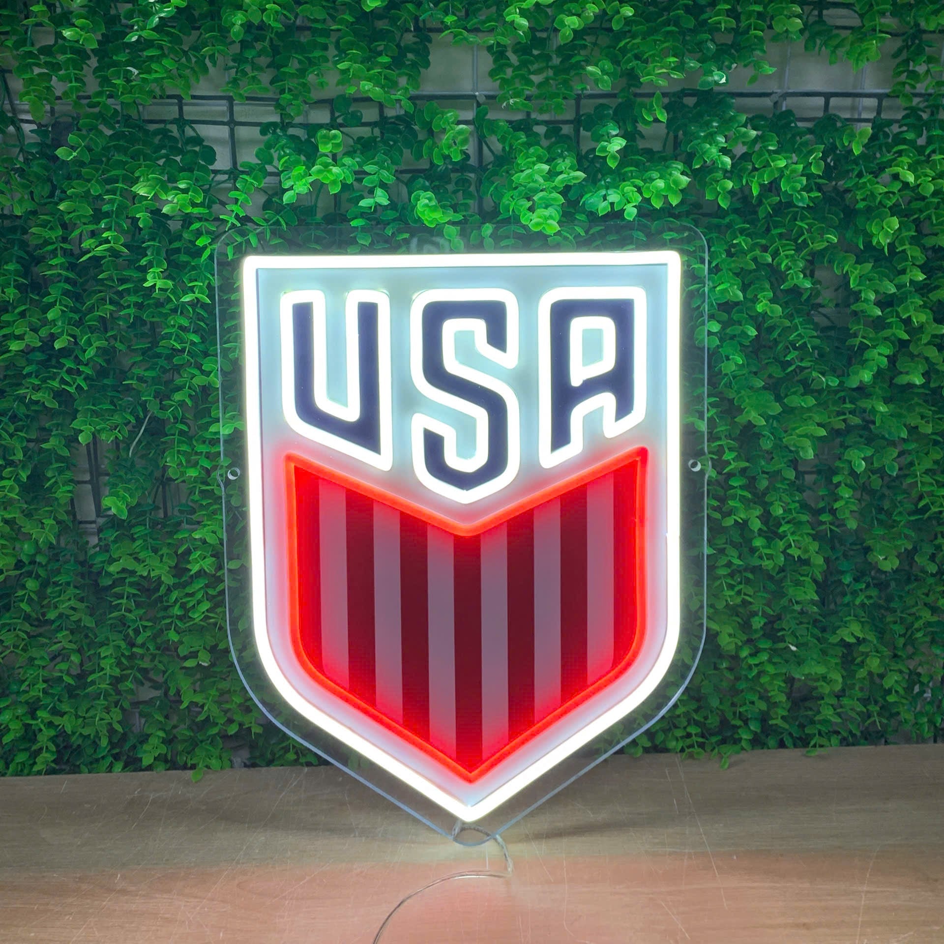 A shield-shaped USA soccer logo sign with a glowing white border, navy blue 'USA' lettering, and red and white vertical stripes, displayed against a green leaf wall.