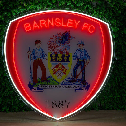 A large illuminated Barnsley FC shield sign mounted against a green leafy wall. The sign features a glowing red and white border, the club name in red neon at the top, a detailed crest in the center, and the year 1887 at the bottom.