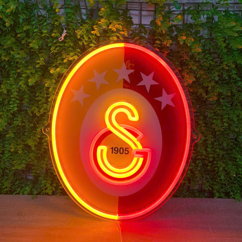 A glowing oval neon sign featuring the Turkish football club Sıvesır logo, with a red and yellow gradient background, white stars, and the year 1905, displayed against a green ivy-covered wall.