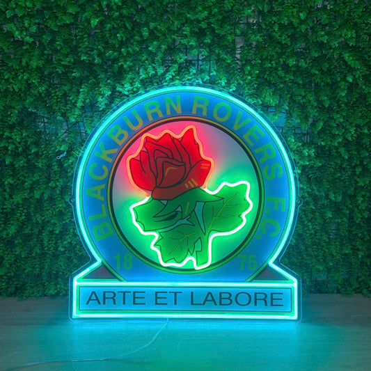 A circular neon-style sign featuring a red rose over green leaves, with the words 'BLACKBURN ROWERS' around the top and 'ARTE ET LABORE' on the base, set against a green artificial hedge wall.